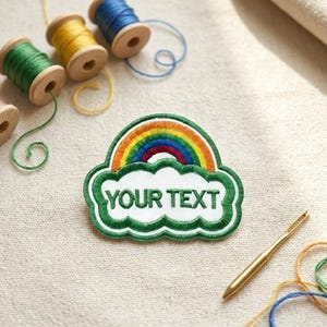 Op de afbeelding: Geborduurde patch met een regenboogontwerp boven een wolkvorm met de tekst "YOUR TEXT" in het wit. De patch is groen omlijnd. Kleurrijke garenklosjes en een naald zijn op de achtergrond.
