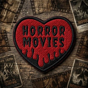 May include: A heart-shaped patch with the words "HORROR MOVIES" in black lettering on a red background. The patch has a black border and a dripping effect at the bottom. The background is a wooden surface with vintage movie posters.
