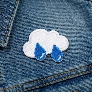 May include: A white embroidered cloud patch with two blue raindrops. The patch is sewn onto a blue denim jacket, with visible stitching and a button.