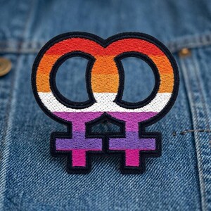May include: Embroidered patch featuring the double Venus symbol, representing lesbian identity. The design incorporates a rainbow gradient of colors, including orange, white, purple, and pink, outlined in black. The patch is affixed to a denim jacket.