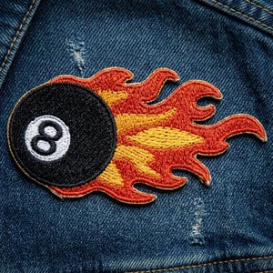 May include: Embroidered patch featuring a black 8-ball with a white number 8, set against a backdrop of stylized flames in shades of red, orange, and yellow. The patch is designed to be ironed on or sewn onto fabric.