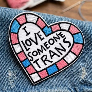 May include: Embroidered heart-shaped patch with the text "I LOVE SOMEONE TRANS" in black. The heart features a white center with the text, and is bordered by pink, blue, and white squares. The patch is on a denim background.