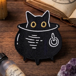 May include: Black cat patch peeking out of a black cauldron with white accents. The cat has yellow eyes. The patch is on a wooden surface with a bottle of Luna Serum and a lit candle in the background.