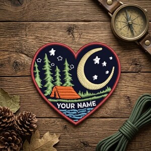 May include: Heart-shaped embroidered patch featuring a camping scene with a tent, trees, moon, and stars. The patch has a red border and includes the text "YOUR NAME". A compass and rope are visible.