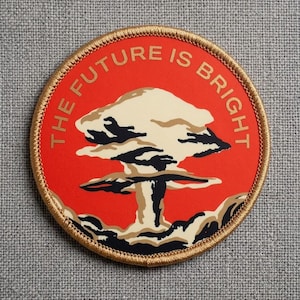 May include: A round patch with a red background and the text "THE FUTURE IS BRIGHT" in gold. The patch features a stylized mushroom cloud design in beige, black, and brown, with a gold embroidered border.