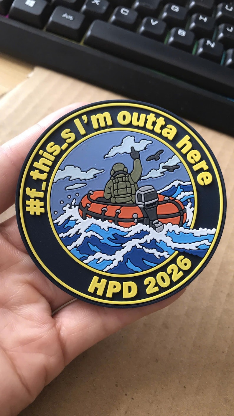 May include: A circular morale patch featuring a cartoon figure in a tactical vest sitting in an orange inflatable boat on blue waves. The text reads #f_this.I'm outta here and HPD 2026 in yellow lettering against a dark blue background.