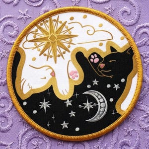 May include: Round embroidered patch featuring a white cat and a black cat in a yin-yang design. The patch has a gold border and includes stars, a crescent moon, and a gold starburst. The cats have pink paw pads and noses.
