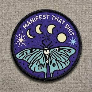 May include: Round patch with a purple background and a black border. The patch features a light blue moth with intricate wing patterns. Above the moth are three crescent moons, stars, and the text "MANIFEST THAT SHIT" in white.