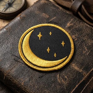 May include: Embroidered patch featuring a crescent moon and four stars on a black background, framed by a gold border. The patch is circular and has a celestial theme. The patch is placed on a textured, brown leather surface.