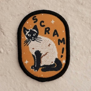 May include: An oval patch featuring a Siamese cat illustration in black and white, set against an orange background. The word "SCRAM!" is written in orange letters around the cat. The patch has a black border and small star accents.