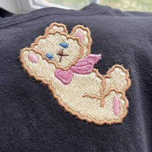 May include: A dark gray sweatshirt with an embroidered teddy bear patch. The teddy bear has a cream-colored body, pink ears and paws, blue eyes, and a pink bow and wings. The patch is outlined in brown.