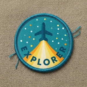 May include: A round, teal patch with the word "EXPLORER" in yellow and orange. A blue airplane is ascending from a yellow and orange light burst against a teal background with white and orange dots. The patch has a teal border.