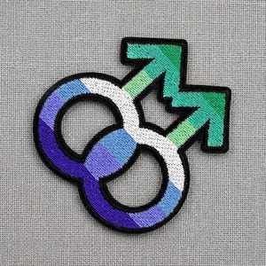 May include: Embroidered patch featuring the intertwined male symbols in a design representing the gay pride flag. The patch has a black border, with the symbols colored in shades of blue, white, and green. The patch is on a gray fabric background.