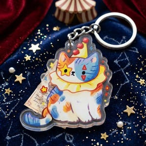 May include: A clear acrylic keychain featuring a cartoon cat dressed as a clown. The cat has orange, white, and blue markings, a star-shaped eye, and a ruffled collar. The keychain has a silver keyring and chain. The background is a dark blue velvet.