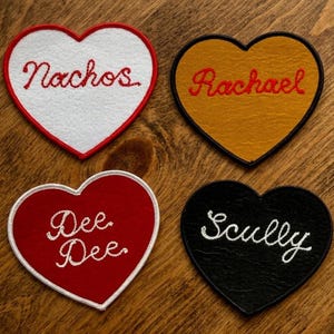 May include: Four heart-shaped patches with embroidered text. One is white with "Nachos" in red, another is gold with "Rachael" in red, a red one with "Dee Dee" in white, and a black one with "Scully" in white.