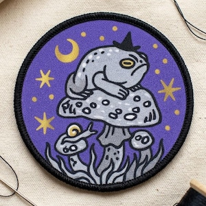 May include: Round patch with a purple background featuring a gray frog wearing a black witch hat, sitting on a mushroom. The patch has gold stars and a crescent moon. A snail and smaller mushrooms are also depicted.