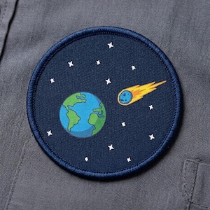 May include: Round embroidered patch featuring a cartoon depiction of Earth, a comet, and stars against a dark blue background. The patch has a navy blue border and is sewn onto a gray fabric.