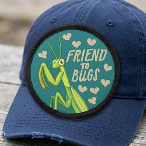 May include: A navy blue baseball cap with a distressed brim. The cap features a round patch with a green praying mantis illustration and the words "FRIEND TO BUGS" in white text, surrounded by small heart shapes.