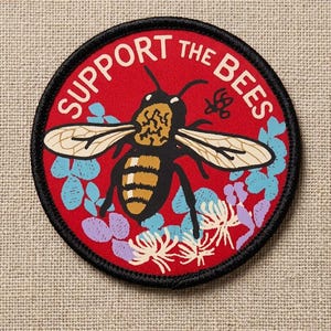 May include: Round patch with a red background and a black border. The patch features a bee illustration with the words "SUPPORT THE BEES" in white. Floral accents in blue, purple, and white surround the bee.