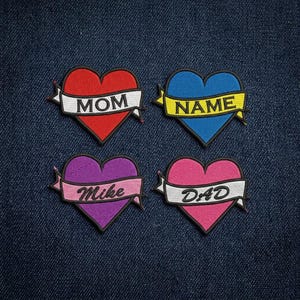 May include: Four embroidered heart patches in red, blue, purple, and pink. Each heart has a banner with text: "MOM", "NAME", "Mike", and "DAD". The patches are on a blue denim background.