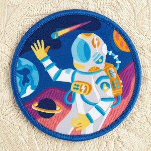 May include: A round patch featuring an astronaut in a white spacesuit, waving in a colorful outer space scene. The patch has a blue border and includes planets, a shooting star, and a purple and pink landscape. The astronaut's suit has yellow accents.