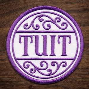 May include: Circular embroidered patch with the word "TUIT" in purple capital letters. The patch has a white center with a purple border and decorative swirls. The patch is on a dark wood surface.