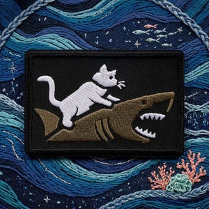 May include: A rectangular black embroidered patch featuring a white cat riding on the back of a brown shark. The shark has an open mouth with visible white teeth. The patch is set against a dark blue background with embroidered ocean waves.