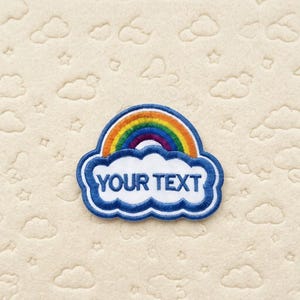 Op de afbeelding: Geborduurde patch met een regenboog boven een wolk met de tekst "YOUR TEXT" in blauw. De regenboog heeft levendige kleuren en de wolk heeft een blauwe omtrek. De patch is geplaatst op een cr&egrave;mekleurige achtergrond met wolken- en sterpatronen.