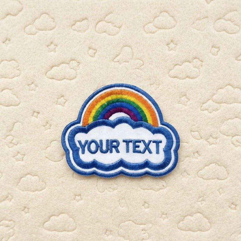 Op de afbeelding: Geborduurde patch met een regenboog boven een wolk met de tekst "YOUR TEXT" in blauw. De regenboog heeft levendige kleuren en de wolk heeft een blauwe omtrek. De patch is geplaatst op een cr&egrave;mekleurige achtergrond met wolken- en sterpatronen.