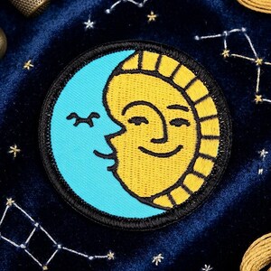 May include: Embroidered patch featuring a smiling sun and moon design. The sun is yellow with a black outline, and the moon is light blue. The patch has a black border and is likely for clothing or accessories.