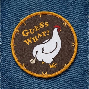 May include: Round embroidered patch with a brown background, a white chicken illustration, and the text "GUESS WHAT?" in yellow. The patch has a gold border and is set against a blue denim background.