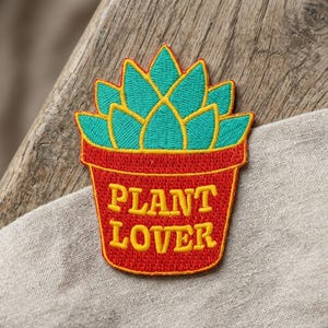 May include: Embroidered patch featuring a teal succulent in a red pot with the words "PLANT LOVER" in yellow. The patch is on a wooden surface and a beige fabric.