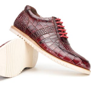 Puede incluir: Zapatos de cuero burdeos con textura de piel de cocodrilo. Los zapatos tienen cordones rojos y suelas blancas. Un zapato está de pie y el otro está tumbado. Sobre un fondo blanco.