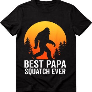 Può includere: T-shirt nera con una silhouette di Bigfoot che cammina davanti a un tramonto giallo e arancione. Il testo "BEST PAPA SQUATCH EVER" è stampato in bianco sotto la grafica. Il design include pini su entrambi i lati di Bigfoot.