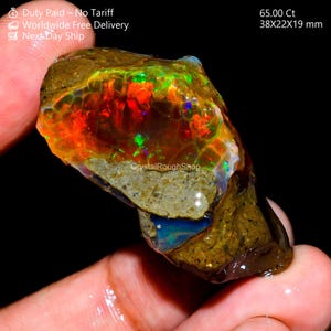 Könnte beinhalten: Ein lebhaftes, roh geschnittenes Opal-Edelstein mit einer Mischung aus Farben, darunter Rot, Grün und Blau. Der Stein wird in einer Hand gehalten und zeigt seine natürliche, unpolierte Textur. Der Opal misst 38x22x19 mm und wiegt 65,00 Ct.