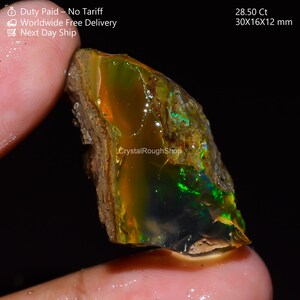 Op de afbeelding: Een ruwe opaal edelsteen met levendige kleuren groen, oranje en geel. De steen meet ongeveer 30x16x12 mm en weegt 28,50 karaat. De tekst "CrystalRoughShop" is zichtbaar.