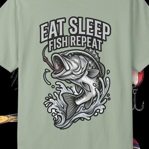 Puede incluir: Camiseta verde claro con el texto "EAT SLEEP FISH REPEAT" y un gráfico de un pez saltando fuera del agua. Cebos y flotadores de pesca están dispuestos alrededor de la camiseta sobre un fondo negro.