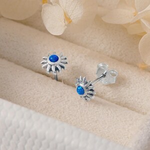 Könnte beinhalten: Ein Paar silberne Daisy-Ohrstecker mit einem blauen Mittelstein. Die Ohrringe werden in einer weißen Schmuckschatulle mit einem weichen, neutralfarbenen Innenraum präsentiert. Getrocknete Blumen sind im Hintergrund.