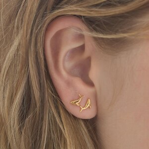 Könnte beinhalten: Nahaufnahme eines Ohrs mit zwei kleinen, goldfarbenen Delfin-Ohrsteckern. Die Ohrringe sind mit Flossen und Schwänzen versehen und befinden sich am Ohrläppchen. Die Person hat blondes Haar.
