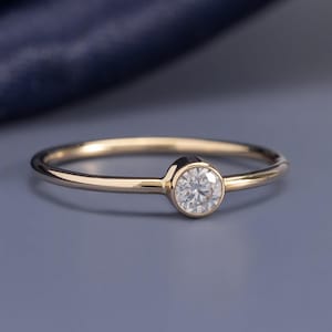 Könnte beinhalten: Ein dünner Goldring mit einem einzelnen runden, klaren, facettierten Edelstein in einer glatten, kreisförmigen Zargenfassung. Die Ringschiene ist poliert und metallisch und zeigt ein schlichtes Design mit einem mittigen Brillantschliff-Stein.