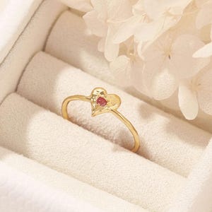 Könnte beinhalten: Ein goldener Ring mit herzförmigem Design und einem kleinen roten Edelstein. Der Ring wird in einer weißen Schmuckschatulle mit weichem, samtigem Innenraum präsentiert. Weiße Blumen sind im Hintergrund.