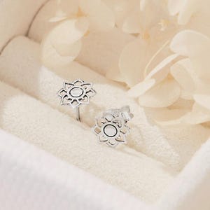 May include: Silver stud earrings with a floral design. Each earring has an openwork pattern around a central white circle. The earrings are displayed in a white jewellery box, with soft, neutral-toned flowers in the background.