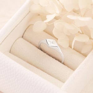 Könnte beinhalten: Ein silberner Ring mit quadratischer Fassung und einem vierblättrigen Design. Der Ring wird in einer weißen Schmuckschatulle mit weichem, strukturiertem Innenraum präsentiert. Getrocknete Blumen sind im Hintergrund.