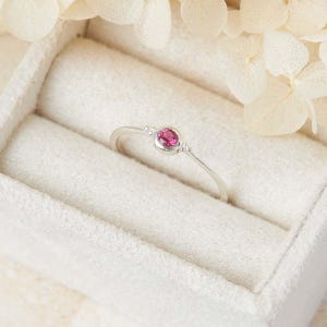 May include: A thin silver ring featuring a single round, faceted pink gemstone set in a bezel setting. The band is slender and metallic, showcasing a minimalist design with the pink stone as the central focal point of the jewelry piece.