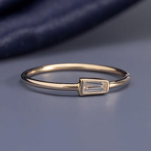 Könnte beinhalten: Ein dünner Goldring mit einem einzelnen, horizontal gefassten, klaren Edelstein im Baguetteschliff. Die Ringschiene ist glatt und poliert, wobei der rechteckige Stein in die Metallfassung integriert ist, was ein schlankes und durchgehendes Profil ergibt.