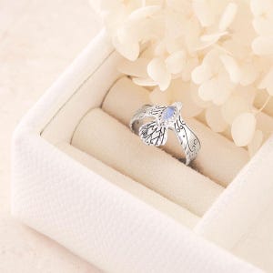 Könnte beinhalten: Ein silberner Ring mit offenem Design, der blattartige Details und einen zentralen, oval geformten, hellblauen Edelstein aufweist. Der Ring wird in einer weißen Schmuckschatulle mit weicher, neutral getönter Beleuchtung präsentiert.