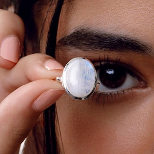 Könnte beinhalten: Ein silberner Ring mit einem ovalen, irisierenden Mondstein. Der Stein hat eine milchig-weiße Basis mit blauen und weißen Mustern. Der Ring wird vor dem Auge gehalten, um die Farbe und das Design des Steins zu zeigen.