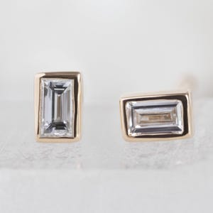 Könnte beinhalten: Zwei goldene Ohrstecker, jeweils mit einem rechteckigen, klaren Edelstein. Die Edelsteine sind in einer goldenen Lünette gefasst. Die Ohrringe werden auf einer weißen Oberfläche präsentiert.