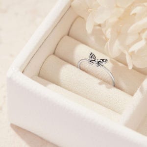 May include: A silver ring with a butterfly design. The butterfly has a detailed pattern and is centered on the ring's band. The ring is displayed in a white jewelry box with a soft, cream-colored interior. Decorative white flowers are in the background.