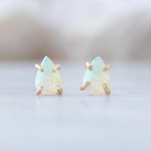 Könnte beinhalten: Ein Paar Ohrstecker mit birnenförmigen Opalen in Goldfassungen. Jeder Opal zeigt eine milchig-weiße Basis mit irisierenden Farbblitzen, gehalten von vier Goldkrallen, die die tropfenförmigen Steine sicher an ihrem Platz fixieren.